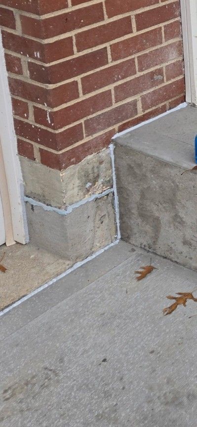 A brick corner of a building with concrete steps and fresh white sealant. Some leaves are on the steps.