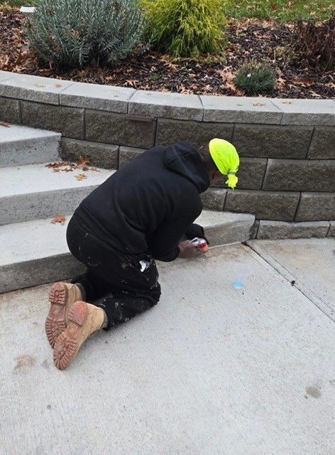 Person kneeling, working on concrete steps. Wears black hoodie, tan work boots, and a neon yellow hat.