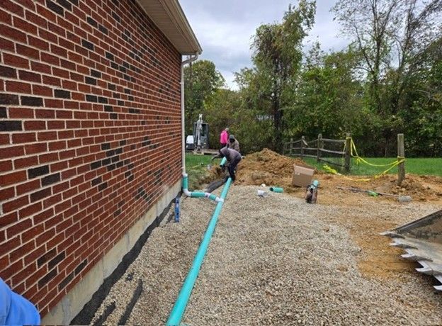 Construction site next to a brick building. Workers install blue pipes in a gravel trench.