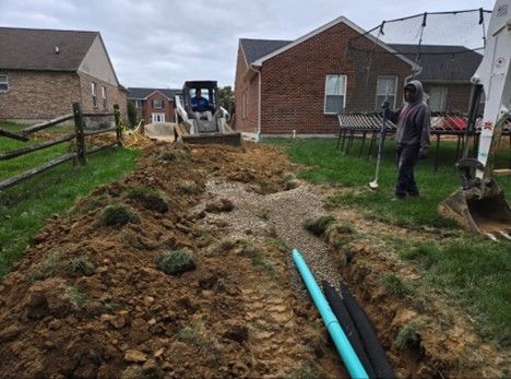Construction site: Trenches dug for drainage pipes near a house. A worker and small earthmover are present.