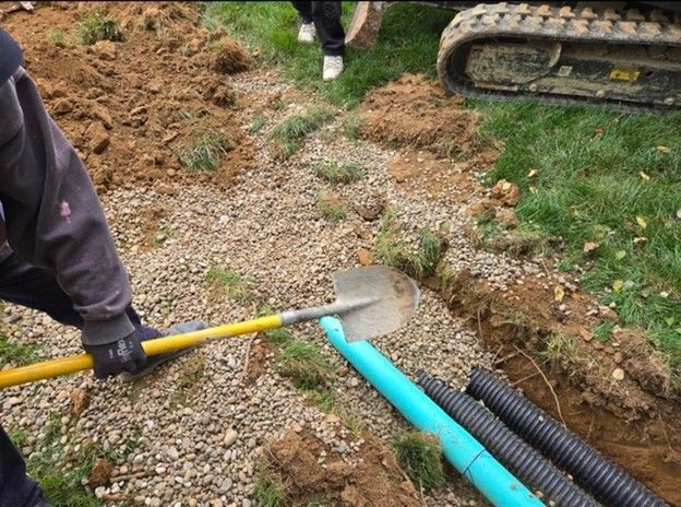 Man shovels gravel around blue and black pipes in a trench next to an excavator and grass.