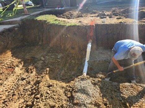 Man digging in a trench near a white pipe, exposed dirt, and a grassy area.