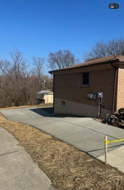 New concrete driveway alongside a brick building and a clearing with trees under a blue sky.