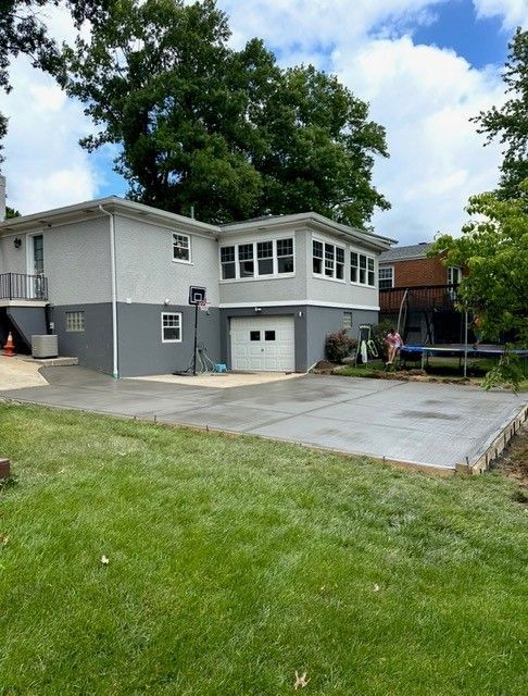Two-story house with light gray upper and darker gray lower exterior. Concrete pad in yard, basketball hoop, trampoline in the background.