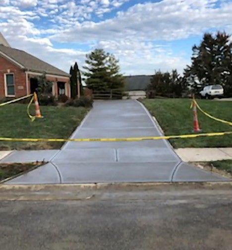Newly poured concrete driveway with yellow caution tape, leading to a brick house on a cloudy day.