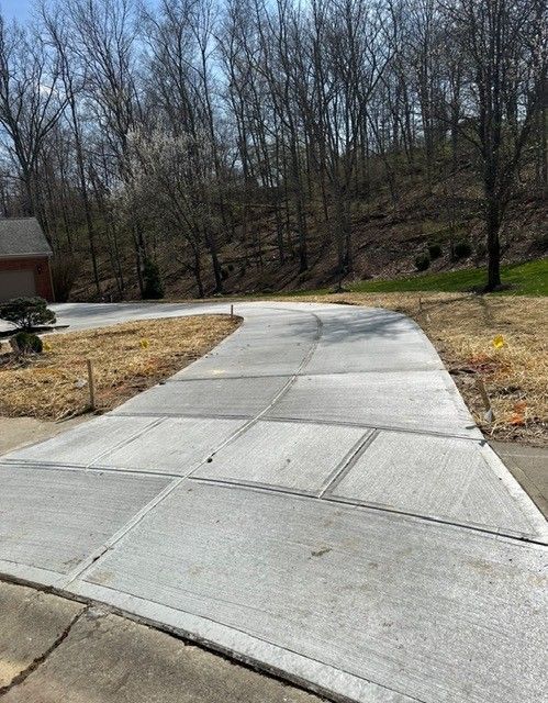 Concrete driveway curving up a slight hill, bordered by dry grass and trees.