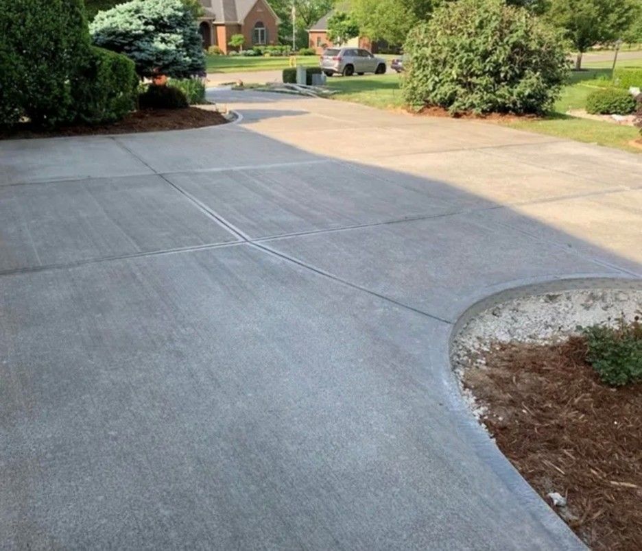 Concrete driveway with expansion joints and curved edge, adjacent to landscaping and houses.