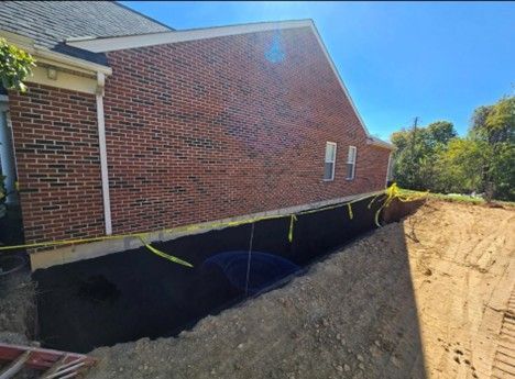A house with a brick exterior has an excavated trench along its foundation. Yellow caution tape is present.