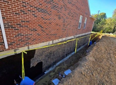 Construction site next to brick building. Men in trench, yellow caution tape, dirt, blue sky.