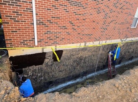 Construction workers near a brick building's foundation. One uses a ladder, the other is in a trench.