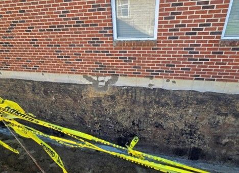 Brick wall foundation with exposed soil and yellow caution tape. Damaged concrete patch near the window.