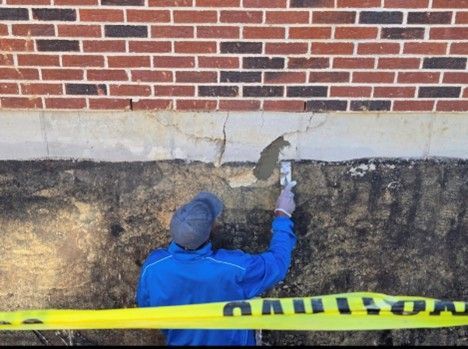 Person repairing a cracked foundation wall with a trowel, brick building in the background, caution tape in the foreground.