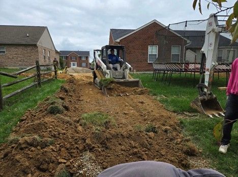 A small excavator digging a trench in a backyard, with a person operating it. Brown dirt and grass present. Houses in background.