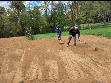 Two people raking soil in an outdoor area, preparing a planting space; trees and fence in background.