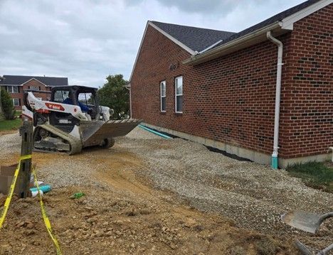 A skid steer compacting gravel next to a brick building. Construction site.