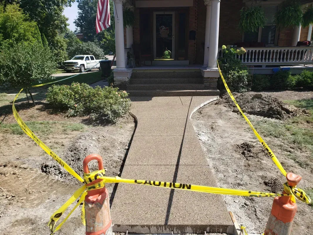 Concrete walkway leading to a house, with yellow caution tape, dirt, and landscaping.