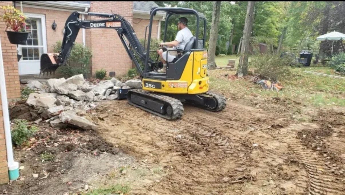 Man operating a yellow mini excavator removing concrete near a house.