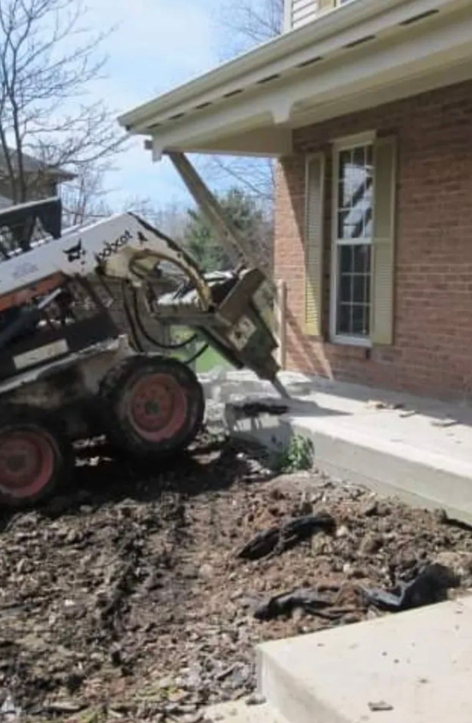Bobcat with jackhammer breaking up concrete next to a house with red brick and a white porch.