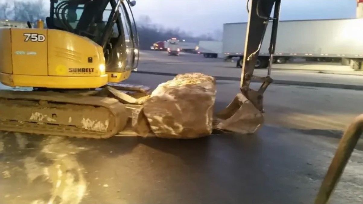 Yellow excavator lifting a large rock at a construction site near trucks and roadway.