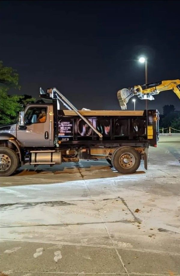 Dump truck being loaded by an excavator in a parking lot at night.