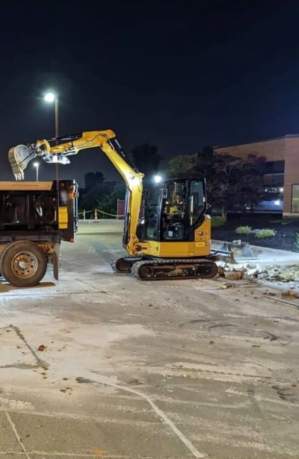 Yellow excavator loading a black dump truck at night. Work lights illuminate a parking lot.