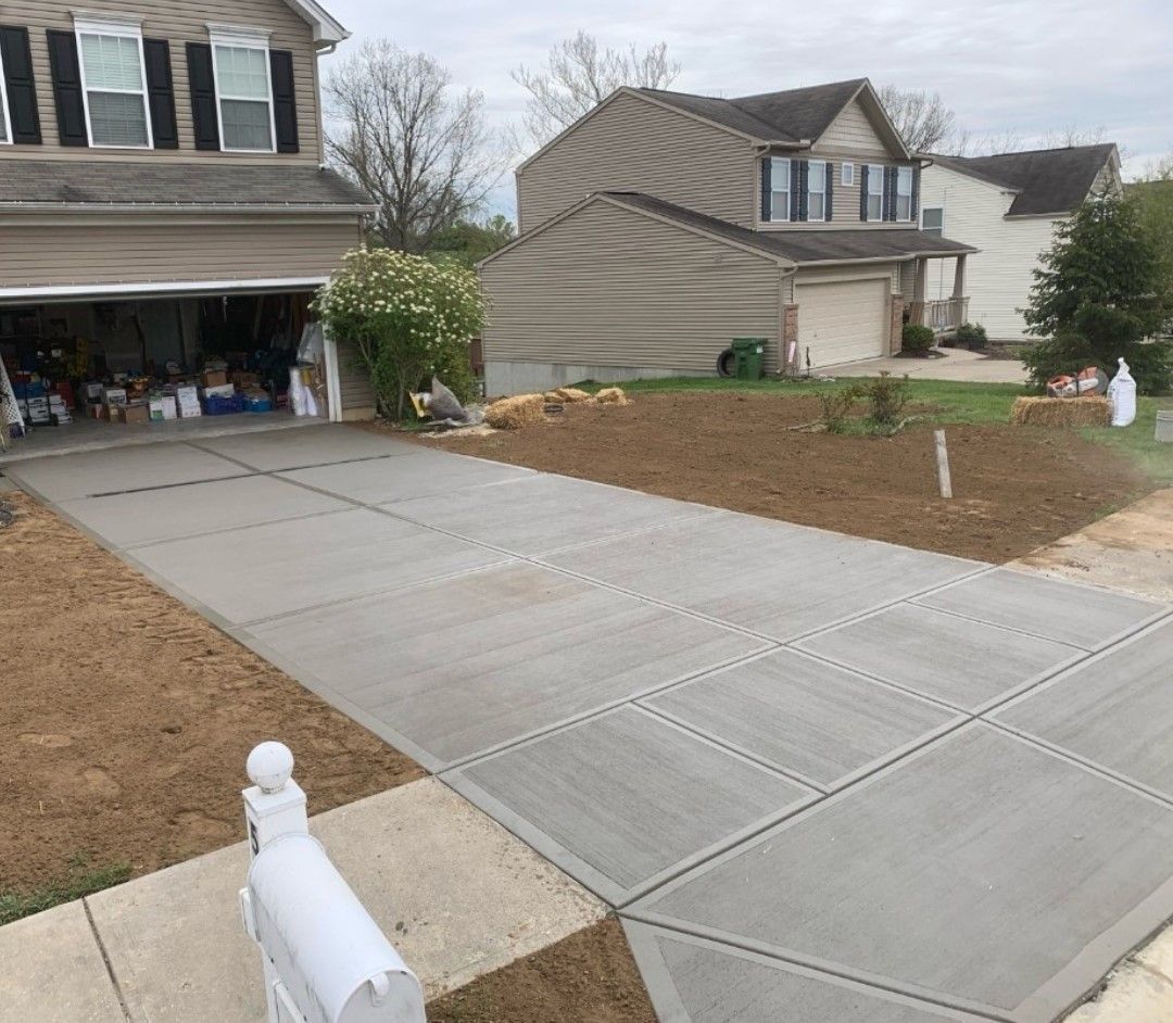 New concrete driveway with adjacent dirt and lawn. Two-story houses in the background.