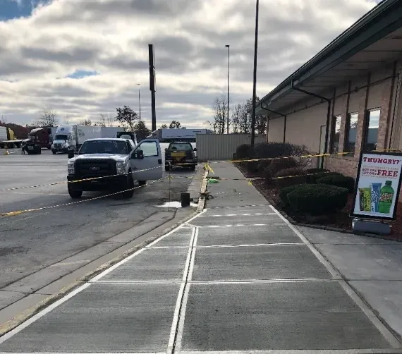Parking lot scene: Trucks, police tape, vehicles, a building with a sign, and a concrete walkway.
