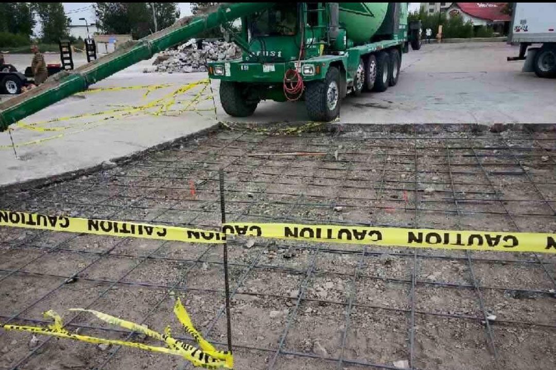 Concrete truck pouring cement over a rebar grid, yellow caution tape on the foreground, outdoors.