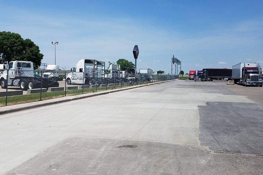 Semi-trucks parked along a concrete lot on a sunny day. Fenced off area.