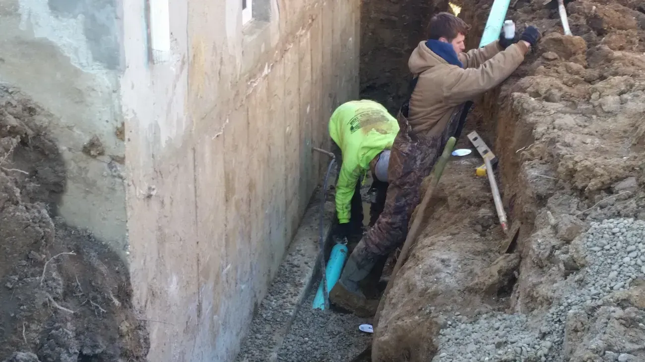 Two workers installing pipe in a narrow trench next to a concrete wall.