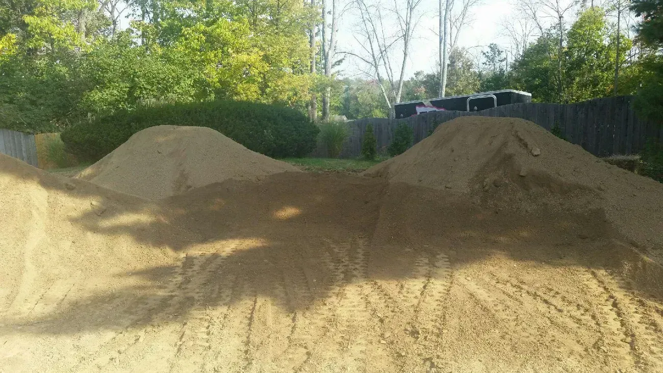 Earthen mounds for a dirt bike jump in a backyard setting, trees in background.