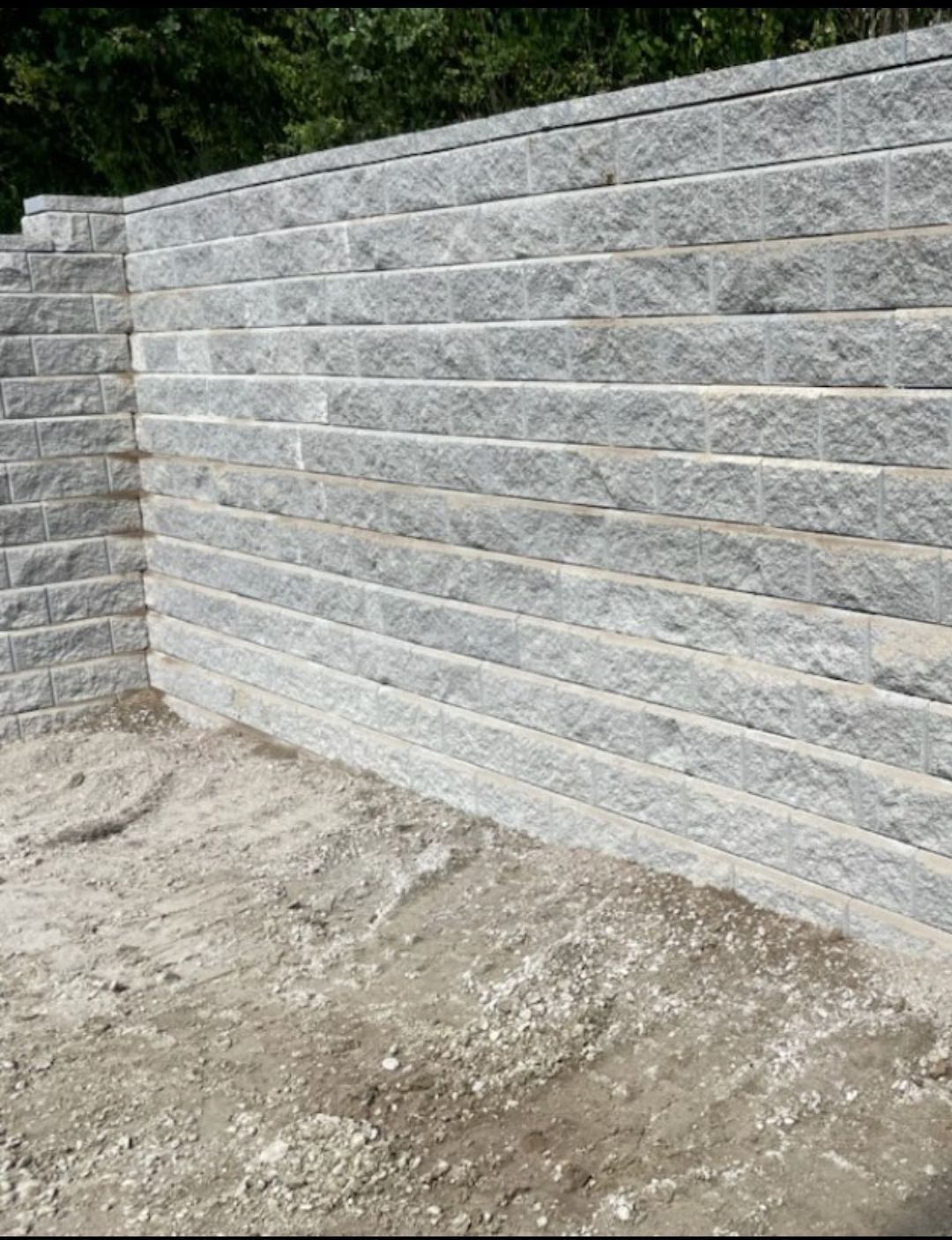 Gray stone retaining wall with horizontal rows, next to a matching pillar. Sandy ground in front.