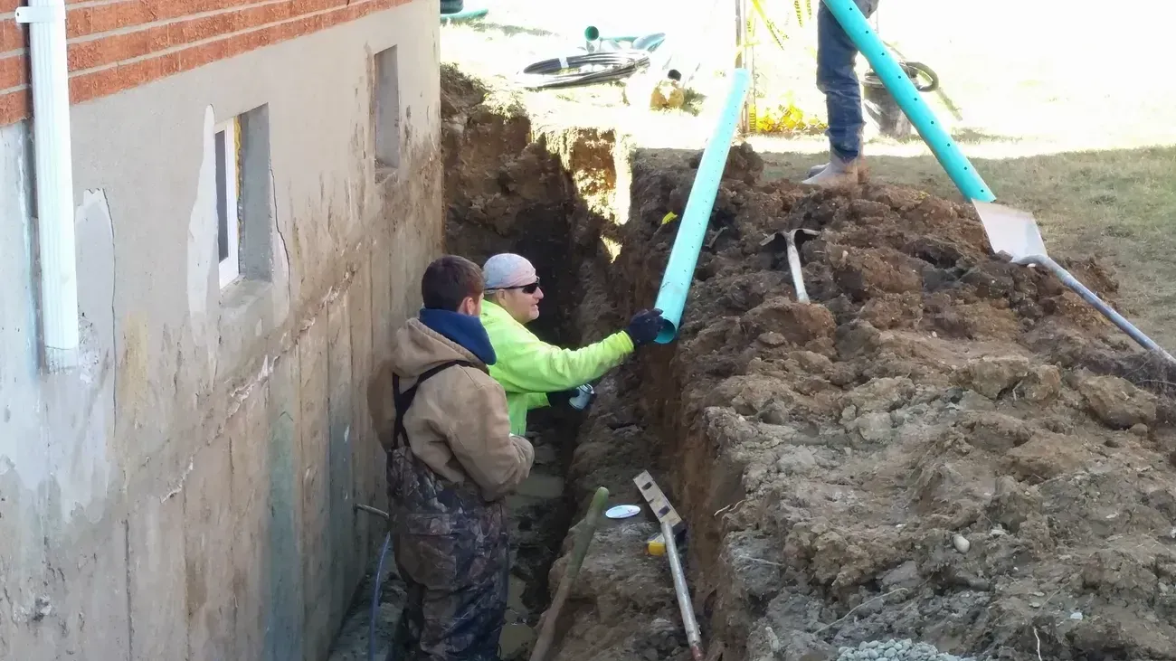 Workers installing a blue pipe near a foundation wall, standing in a trench.