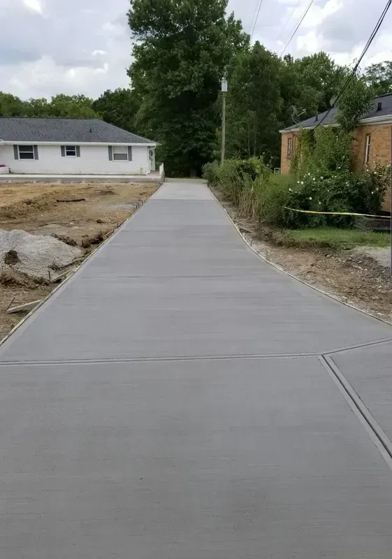 Newly poured concrete driveway between houses, leading toward a wooded area. Gray, overcast sky.