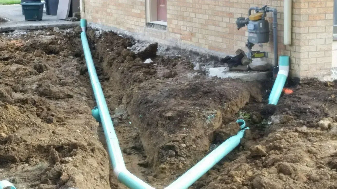 Trench with turquoise pipes along a brick building. Earth surrounds the pipes, construction in progress.