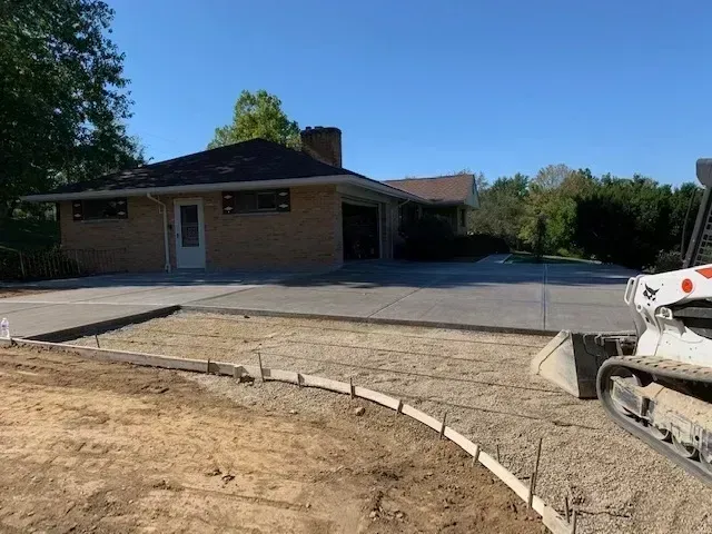 Construction site: new concrete driveway being poured. Brick house with existing driveway in the background.