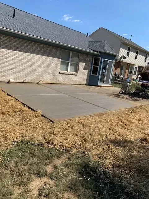 Newly poured concrete patio next to a brick house with a blue door, surrounded by dry grass.