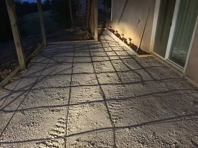 Concrete patio under construction; rebar grid laid on gravel base, adjacent to house with sliding glass door.