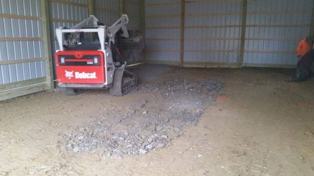 Bobcat skid-steer loader working inside a metal-walled building, moving gravel on a dirt floor.