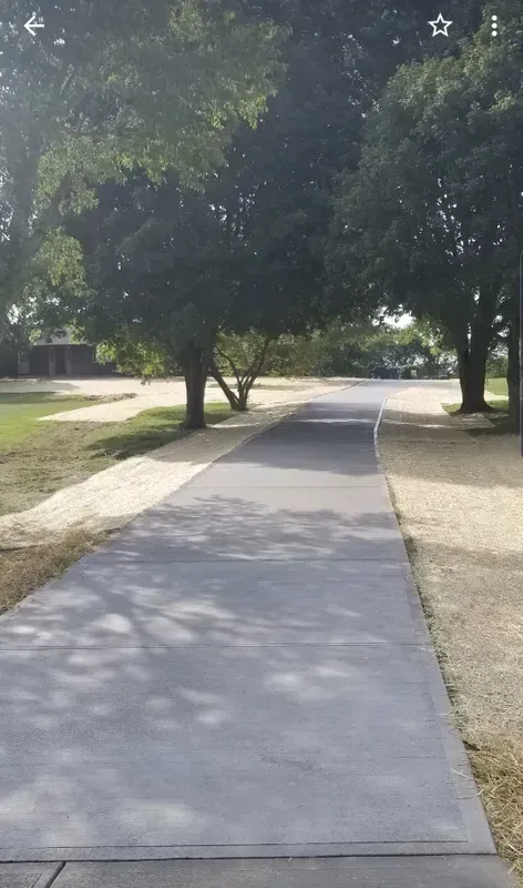 Gray paved path through park, lined with trees and gravel.