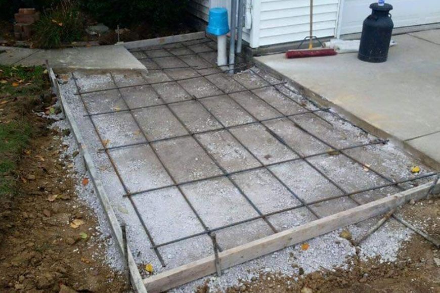 Concrete slab construction in progress, showing rebar grid, wooden form, gravel base, and adjacent existing walkway.