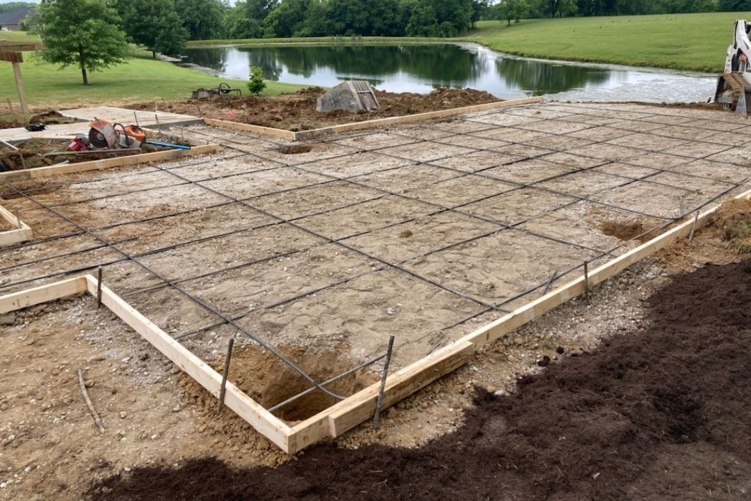Preparation for concrete slab: Wooden forms with rebar grid laid on dirt, near a pond.
