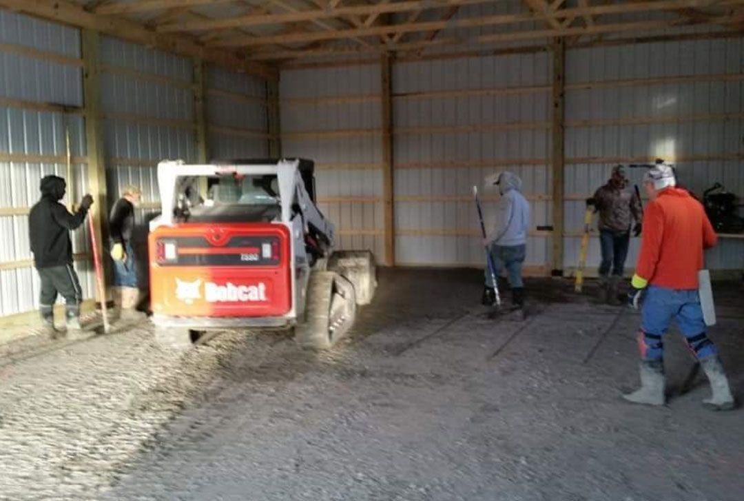 People using tools, including a Bobcat, to work on a gravel floor inside a barn.