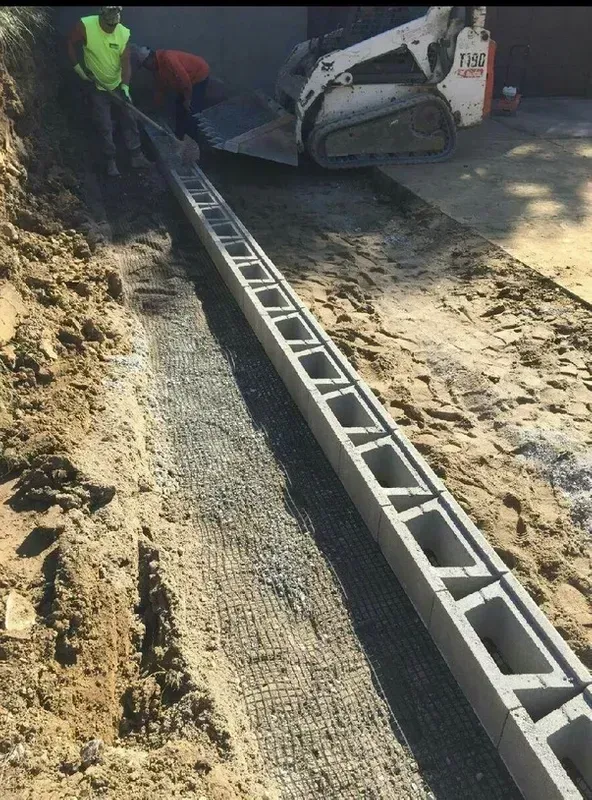 Construction workers laying concrete blocks in a trench filled with gravel; a Bobcat loader is nearby.