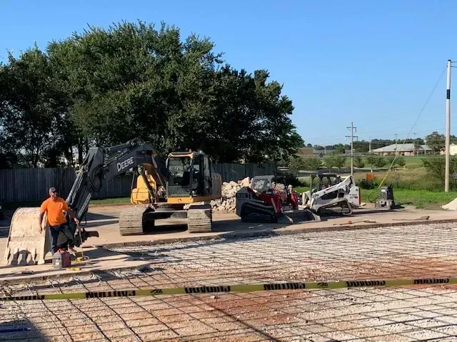 Construction site: Worker, heavy machinery (excavator, skid steer), rebar grid, yellow caution tape, blue sky.