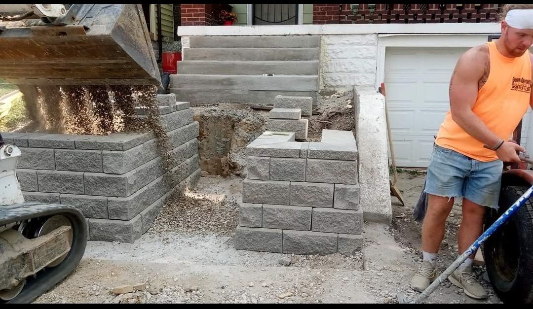 Construction site: man in orange shirt builds retaining wall for steps. Excavator and gray blocks present.