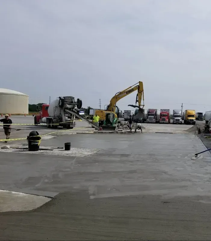 Concrete pour at construction site; cement truck, excavator, workers, trucks in background.