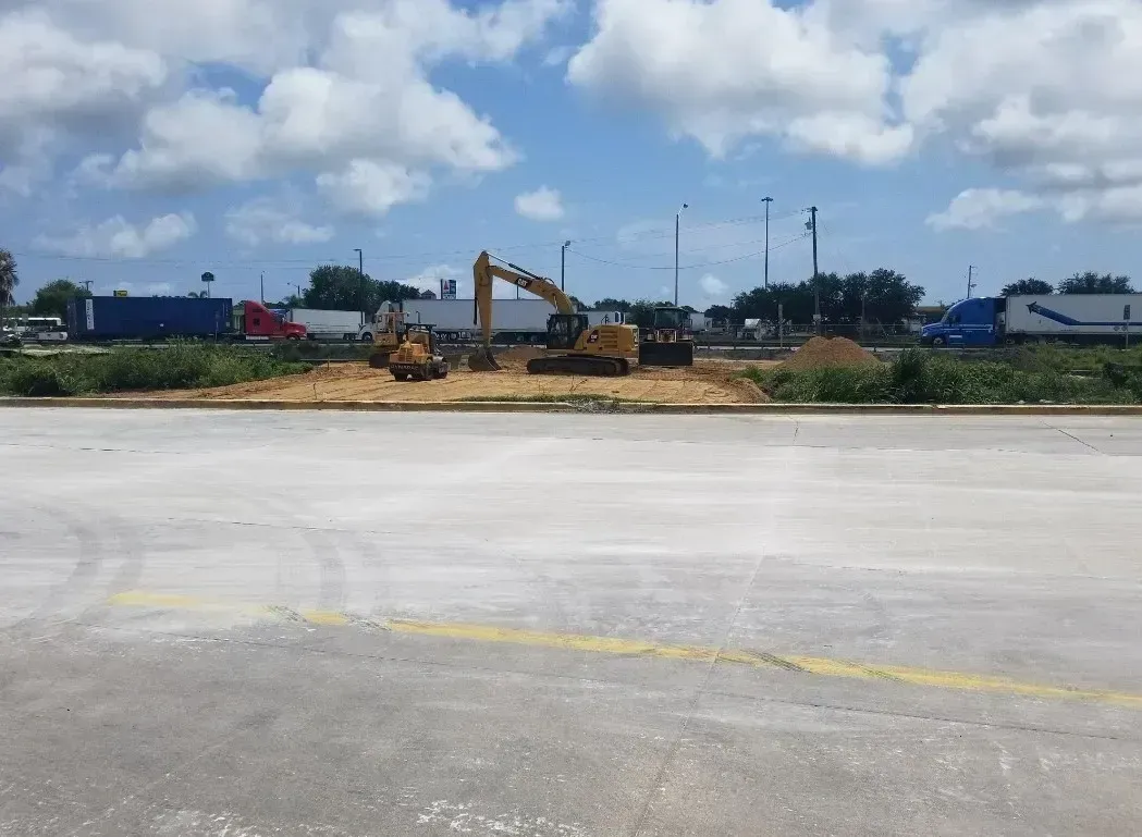 An excavator working on a dirt pile, with trucks and sky in the background.