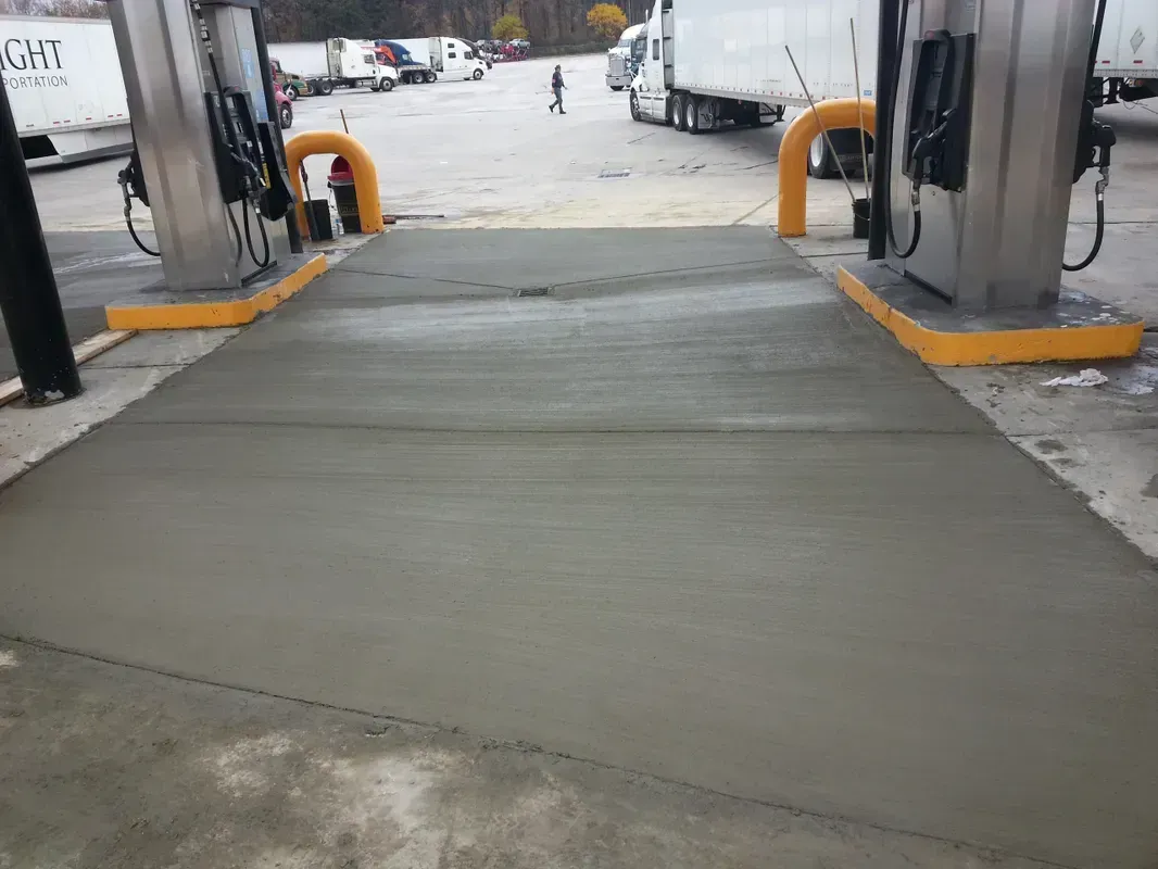New concrete pad at a gas station fueling island, with yellow safety bumpers and fuel pumps.