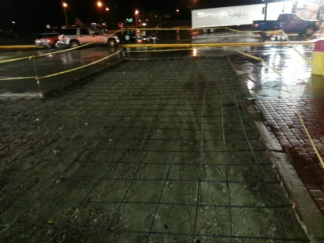 Construction site: metal rebar grid on a concrete pad, surrounded by caution tape. Wet ground, night setting.